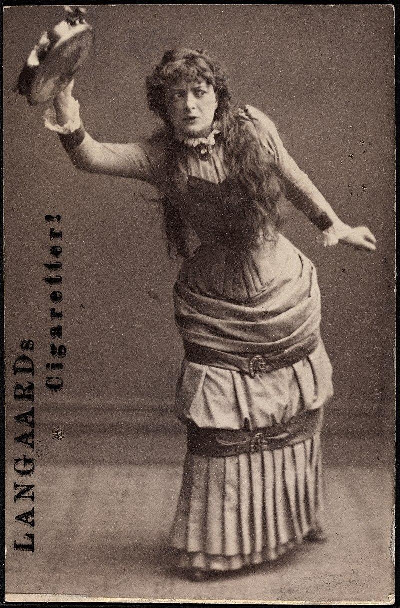 Adeleide Johannessen in character as Nora, from a cigarette card of c. 1880 – c. 1882.

Adeleide Johannessen is captured mid-action, holding a tambourine in her raised hand, evoking an energetic or defiant moment from her performance as Nora.

Her body language is dynamic, and her intense facial expression reflects the emotional depth and complexity of Nora's character, perhaps hinting at her journey of self-discovery and rebellion.

Johannessen wears a full-length Victorian-era dress, with detailed pleating, fitted bodice, and layered skirts that cinch at the waist with a decorative belt.