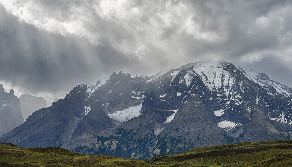 Sunbeams break through the clouds to highlight portions of a towering, snow-capped Patagonian mountain range. In the foreground, a tranquil green plain sets a stark contrast to the rugged peaks above.