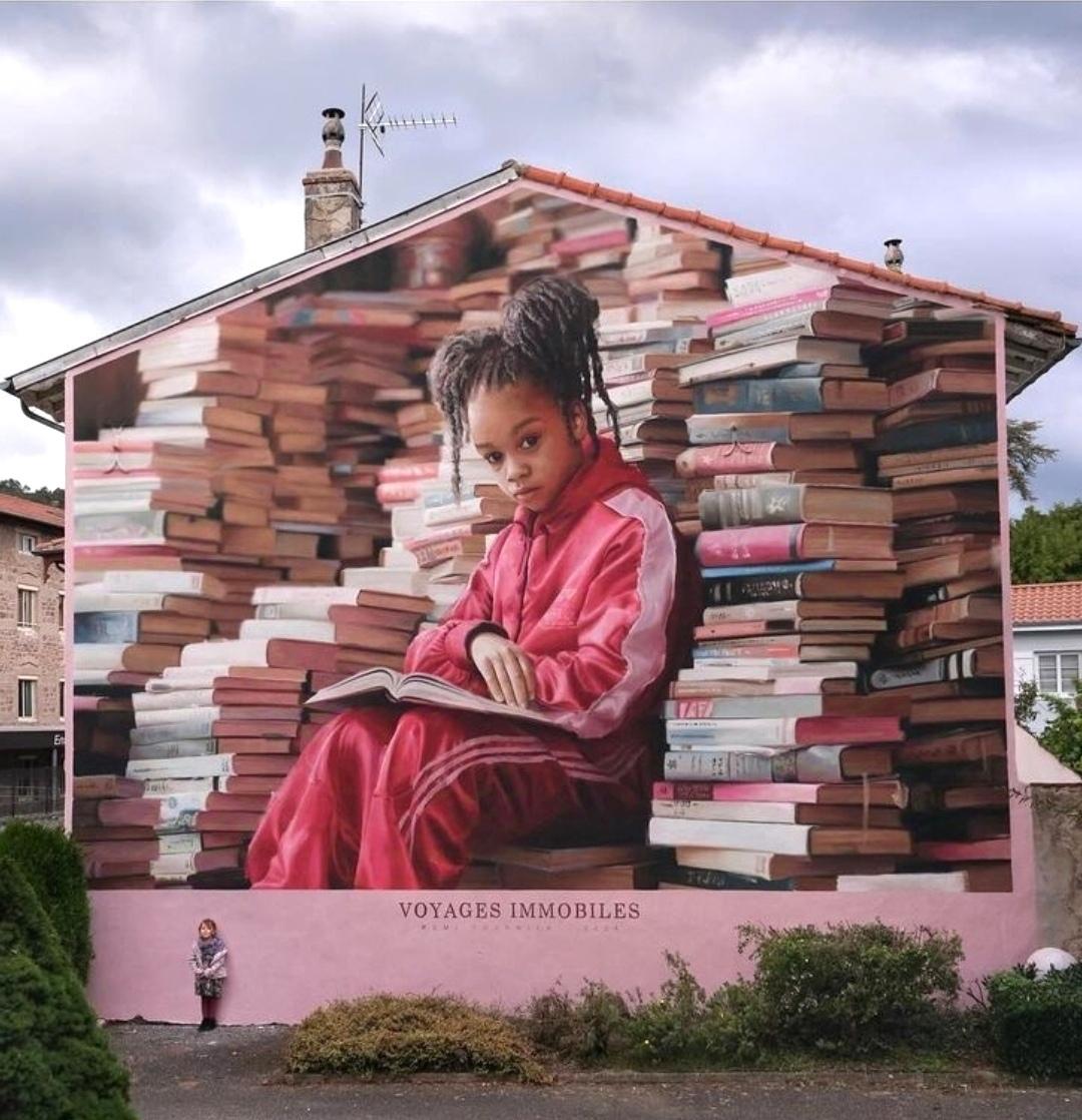 Streetartwall. An impressively realistic, large (10x9 meters) mural was painted on the outside wall of a two-storey building. It shows a little black girl among books. The background and main color is a light pink. The little girl with short hair and small pigtails is sitting with a book on her knees in front of a wall of countless stacked books. She is wearing a pink tracksuit and has briefly looked up at the viewer. The title of the mural is under "Voyages Immobiles" and refers to the background of the picture. Traveling with our thoughts, which know no boundaries thanks to the books.
(The photo shows another little girl standing in front of the wall.)